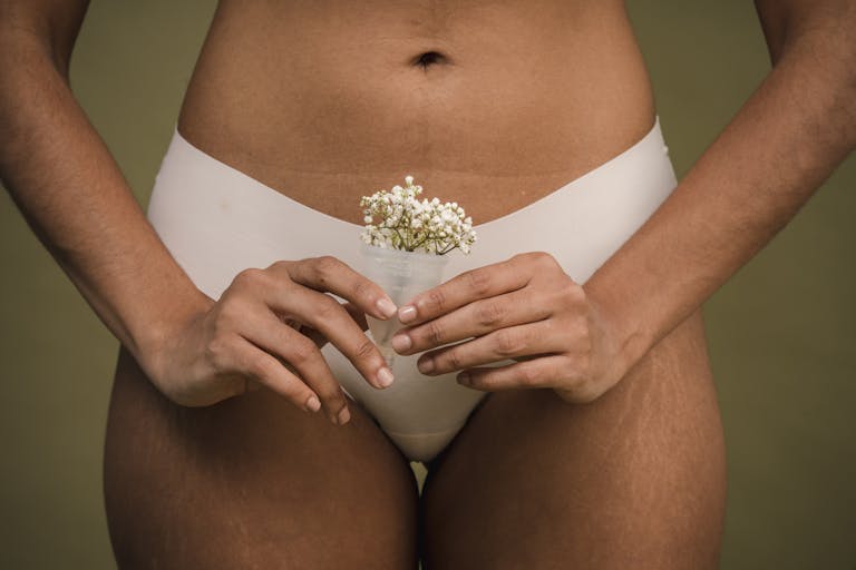 Crop unrecognizable female with stretch marks on hips in white underwear holding menstrual cup with delicate flowers in hands against green background